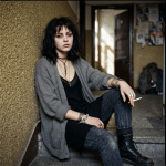 Young woman with dark hair, jewelry, and boots smoking in a hallway